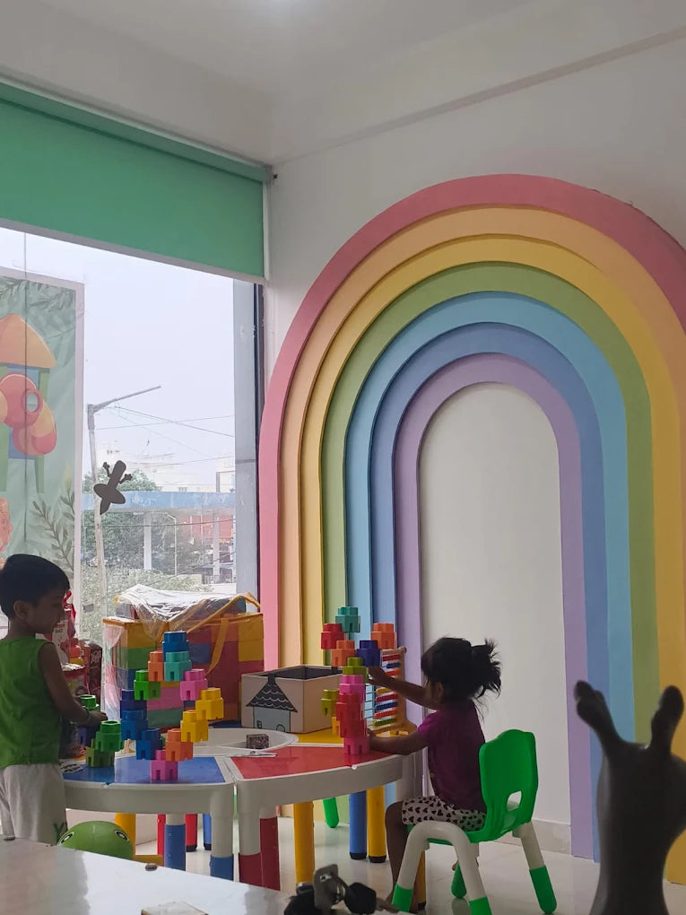 Two young children play with colorful blocks at a table in a brightly decorated room with a rainbow arch.