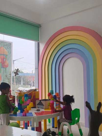 Two young children play with colorful blocks at a table in a brightly decorated room with a rainbow arch.