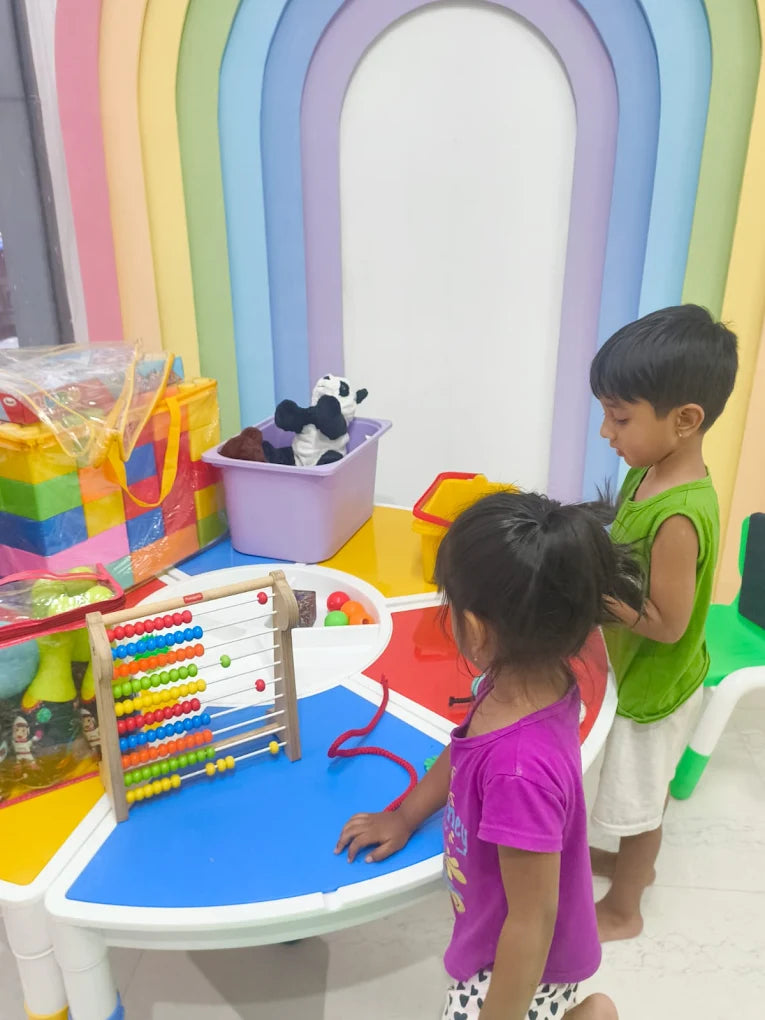 Two young children play with an abacus and toys at a colorful table in front of a rainbow-themed wall.