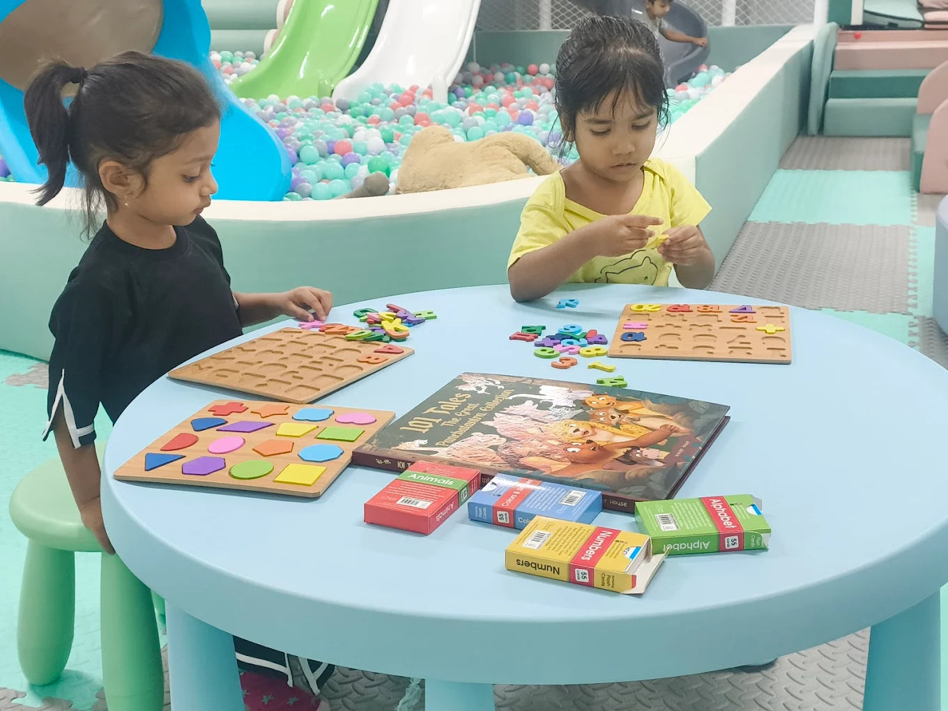 Two young girls play with educational puzzles and cards at a light blue table in a colorful indoor play area.