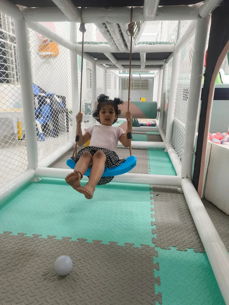 A young child swings on a blue swing in a soft play area with padded floors.
