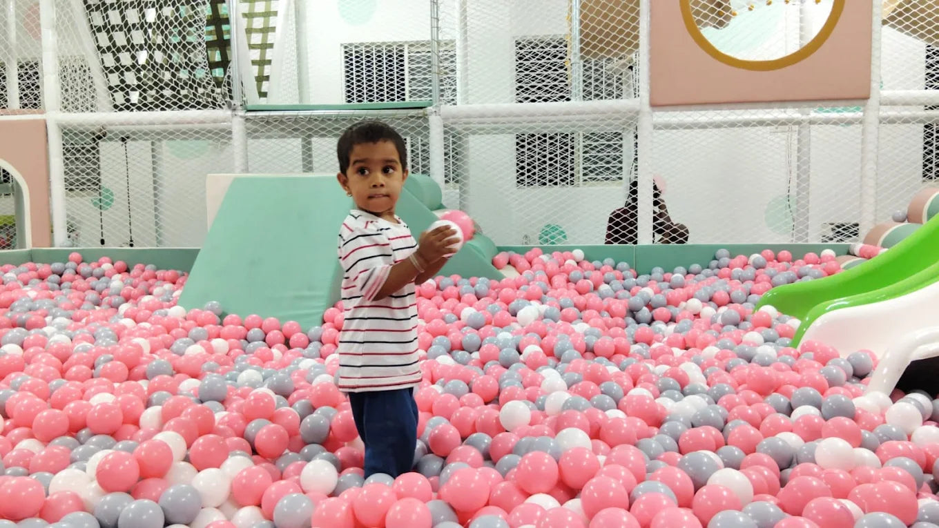 A young child stands in a ball pit filled with pink, gray, and white balls, holding a pink ball.