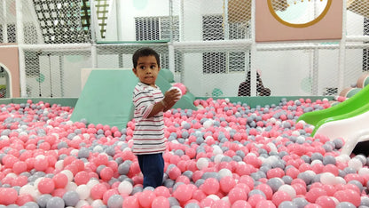 A young child stands in a ball pit filled with pink, gray, and white balls, holding a pink ball.