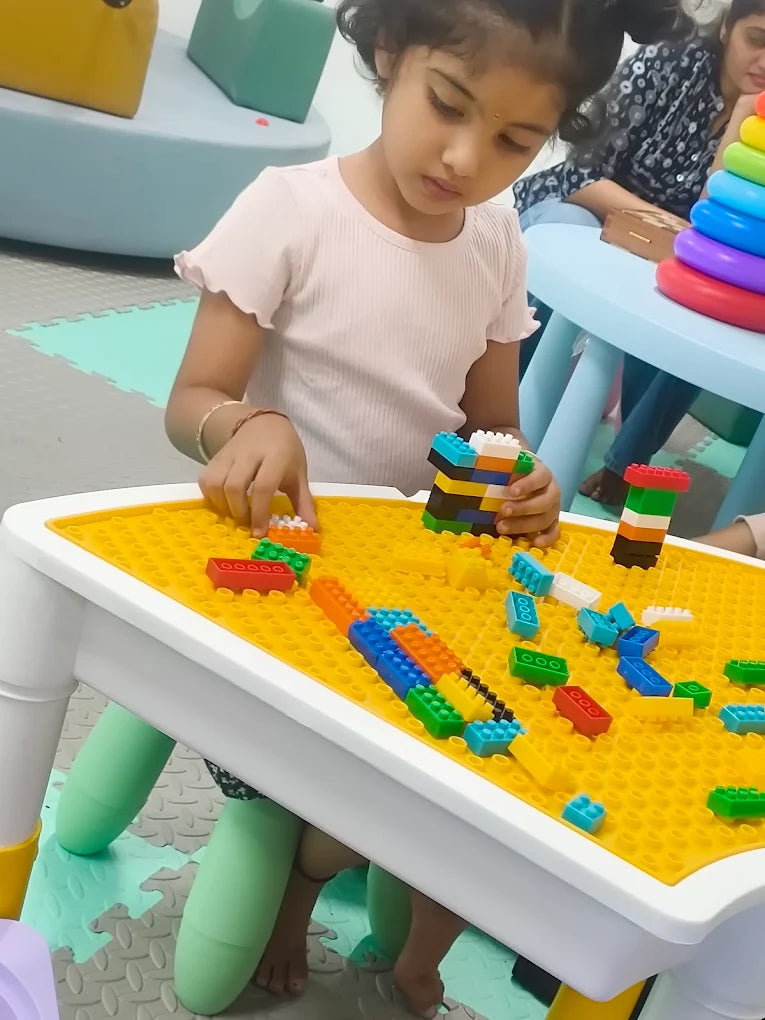 A young girl plays with colorful building blocks on a yellow table.