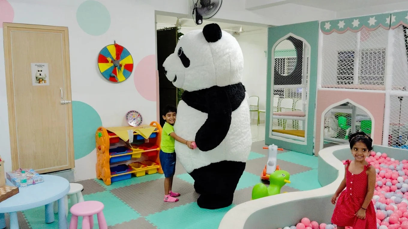 A young boy hugs a giant panda mascot in a colorful playroom, while a young girl smiles in a ball pit.