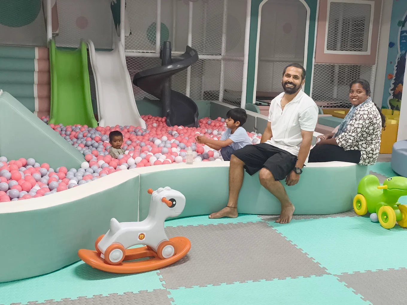 A family enjoys a ball pit at an indoor playground, with a toddler in the balls and adults sitting nearby.