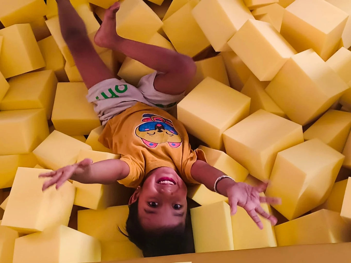A young child playfully tumbles in a pit of yellow foam cubes.