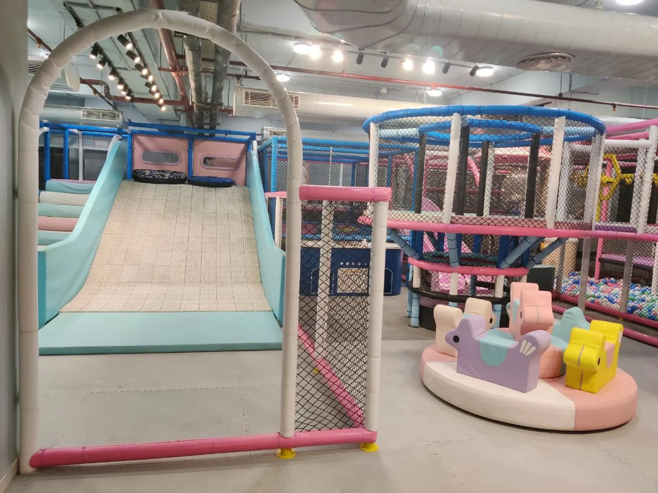 Indoor play area with soft slide, pastel-colored animal rides, and netted climbing structures in a kids entertainment zone