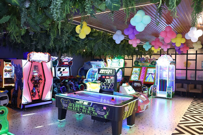 Bright indoor arcade space with glowing air hockey table, racing simulators, and various interactive game machines under leafy ceiling décor