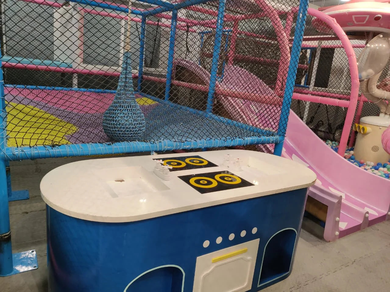 Indoor kids play zone with blue climbing frame, hanging net element, pink slide, and colorful ball pit in safe environment