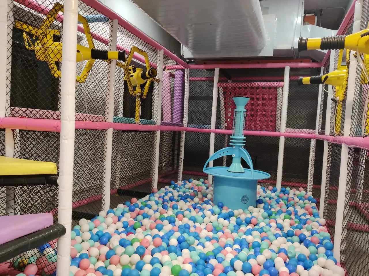 Indoor play zone featuring a ball pit with pastel balls, netted climbing areas, and interactive yellow play cannons
