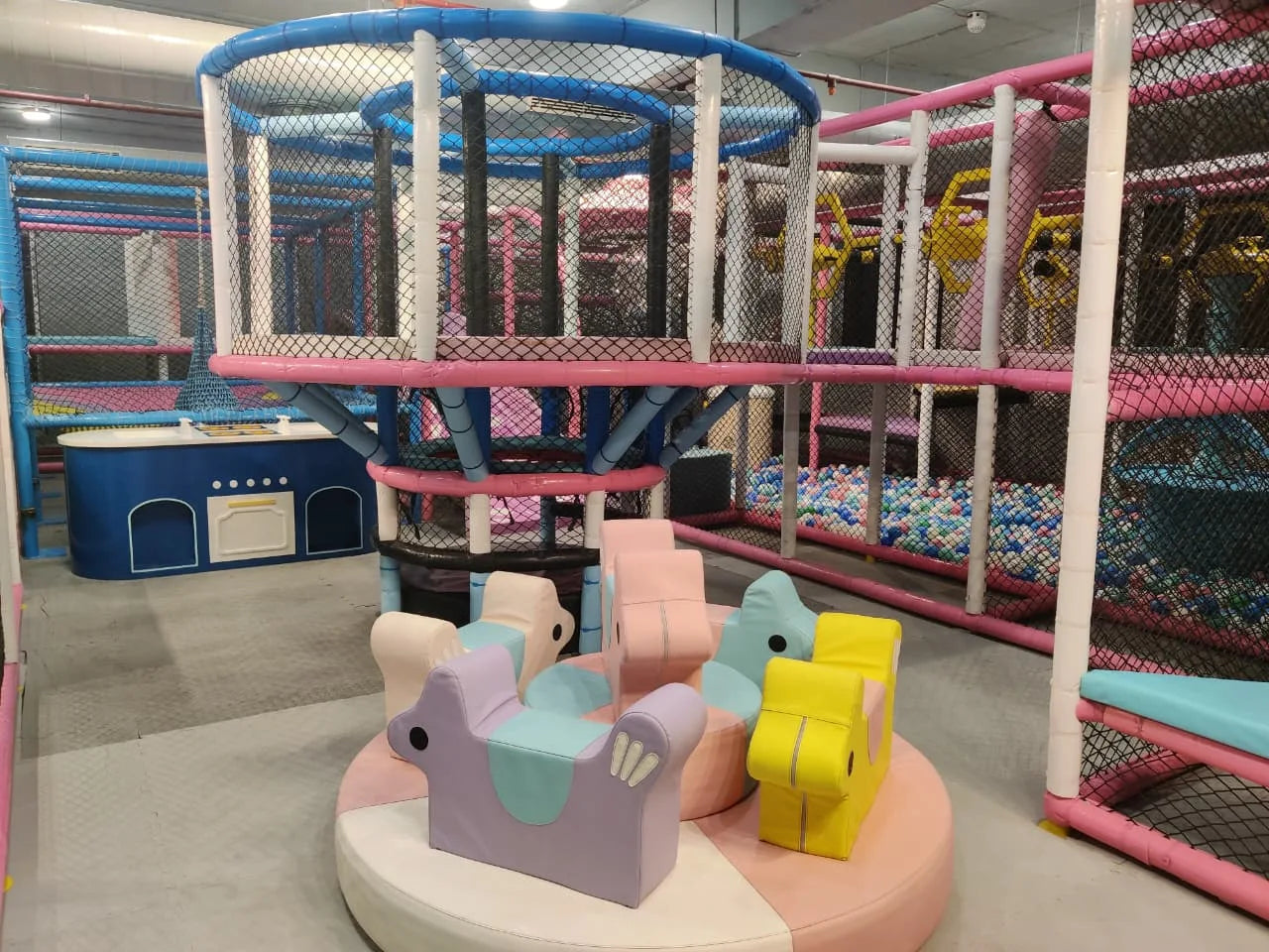 Indoor soft play area with pastel animal-shaped seats and multi-level climbing structures surrounded by safety netting