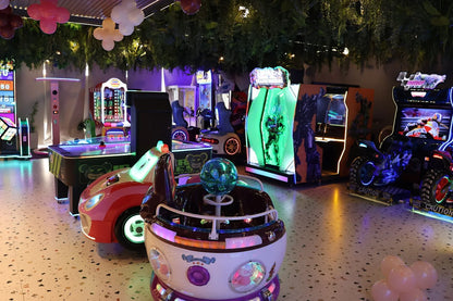 Dimly lit arcade room with glowing ride-on cars, interactive game machines, and leafy ceiling decorations