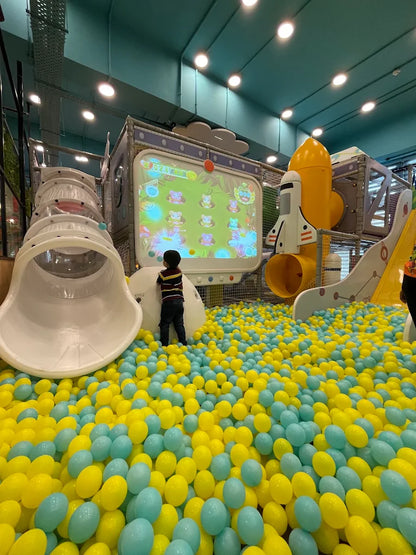 A child plays in a ball pit in an indoor playground with a large screen displaying a game.