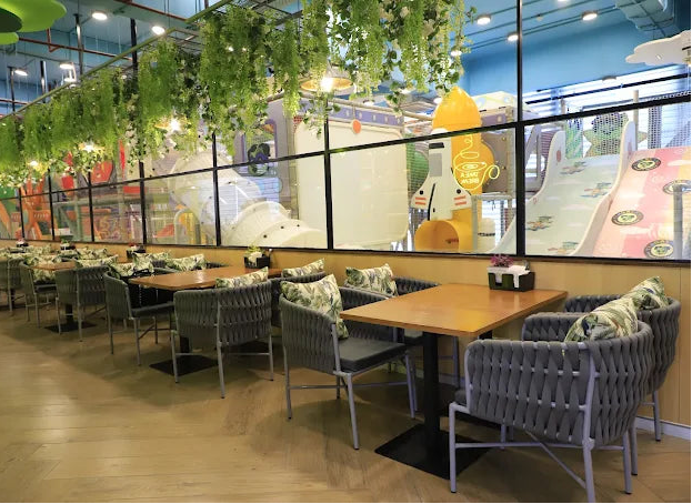 Cafeteria with tables, chairs, and decorative plants on a glass wall. Kids Indoor Play Area visible through the glass wall.