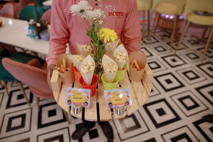 Person holding wooden tray with colorful cups of popcorn and small yellow flowers on patterned floor background