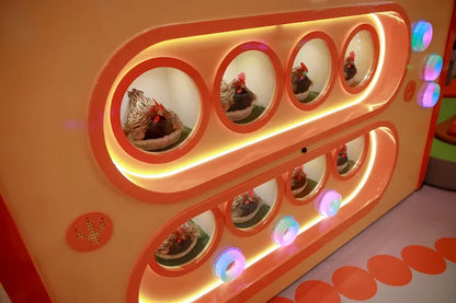 Curved orange play panel with illuminated circular windows showcasing toy chicken figures in a vibrant kids’ zone