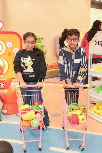 Two children smiling and pushing pink toy shopping carts filled with play fruits in a colorful indoor play area