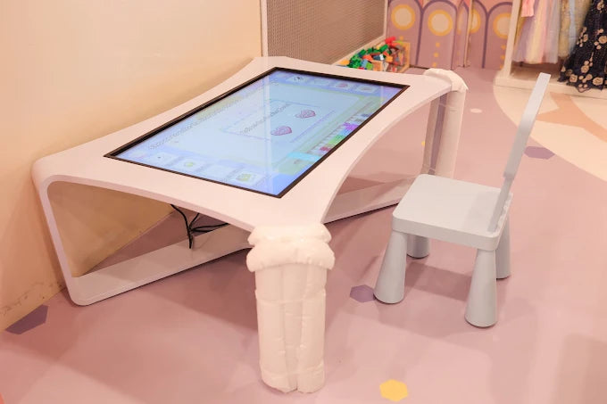 Interactive digital play table with a small light blue chair, set on a pastel pink floor in a kids’ area