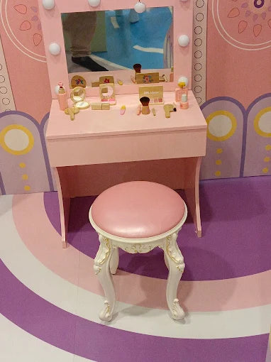 Child-sized pink vanity table with light bulbs and matching cushioned stool in a playful indoor setting