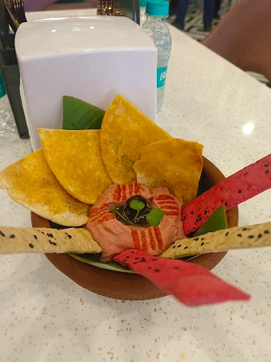 Bowl of colorful chips and creamy dip garnished with herbs served on a green leaf on white speckled surface