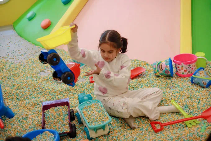 Girl playing with toy trucks and colorful pebbles in a lively indoor kids’ play area at Tooney Tales Gurugram