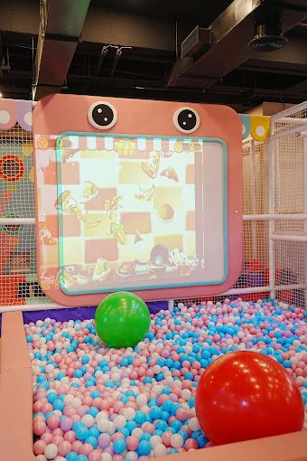Indoor ball pit with blue, pink, and white balls and interactive screen framed by orange panel with eyes