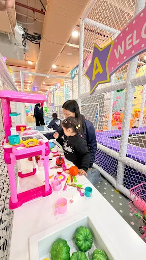 A woman and two young children play with a toy kitchen in a colorful indoor playground.