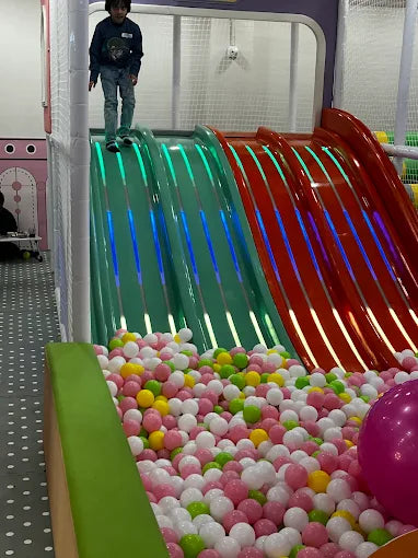 A child stands at the top of a colorful slide at an indoor playground, ready to slide into a ball pit.