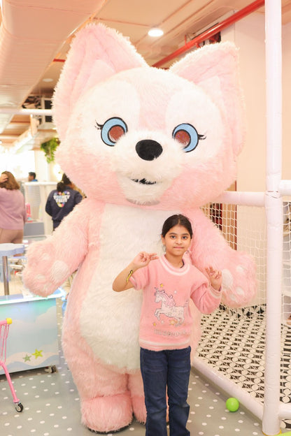 A young girl poses with a large pink mascot in a playful indoor setting.