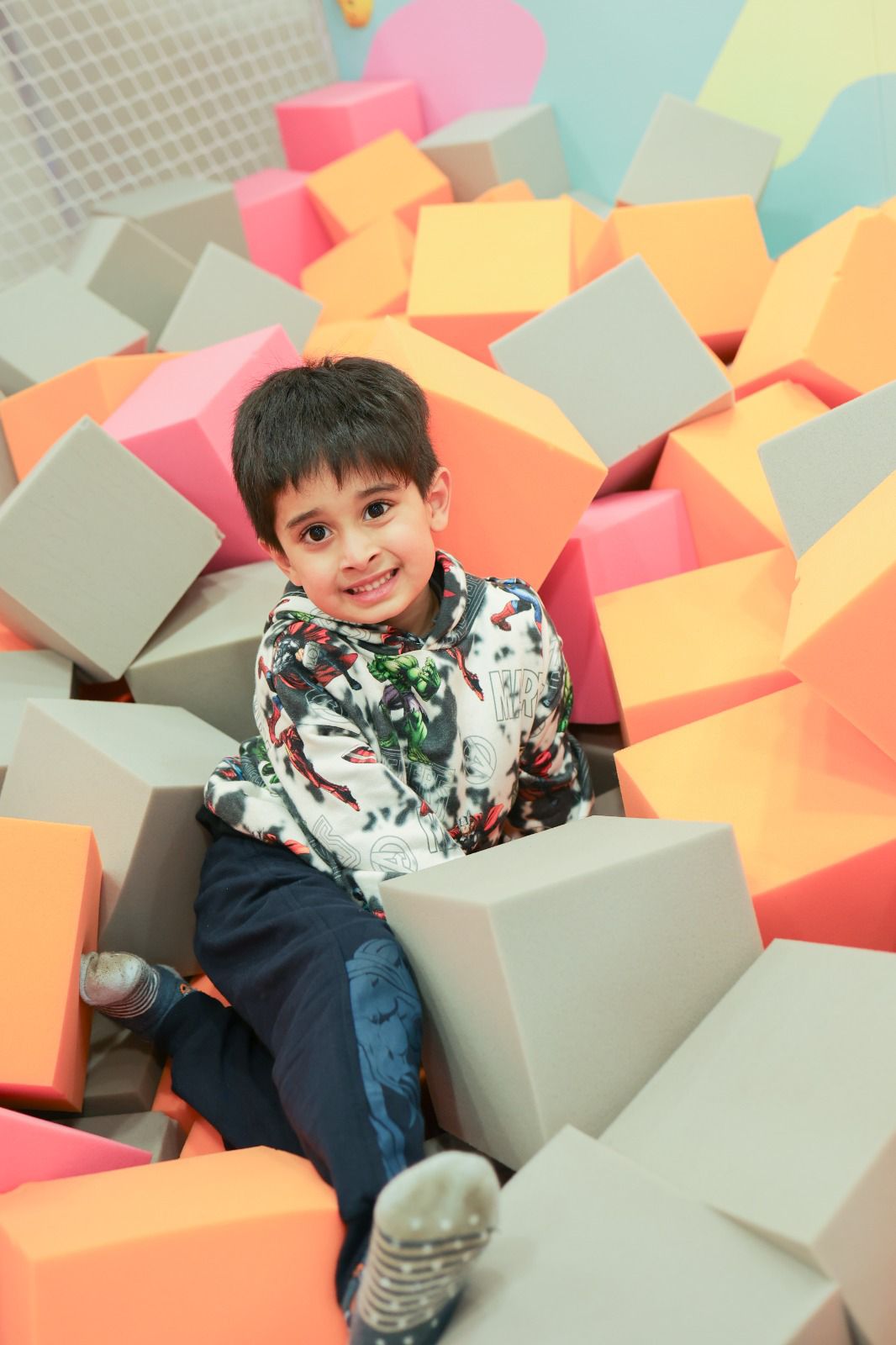 A young boy smiles while sitting in a ball pit filled with colorful foam blocks.