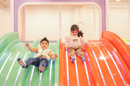 Two young children slide down colorful playground slides, one on green and one on orange.