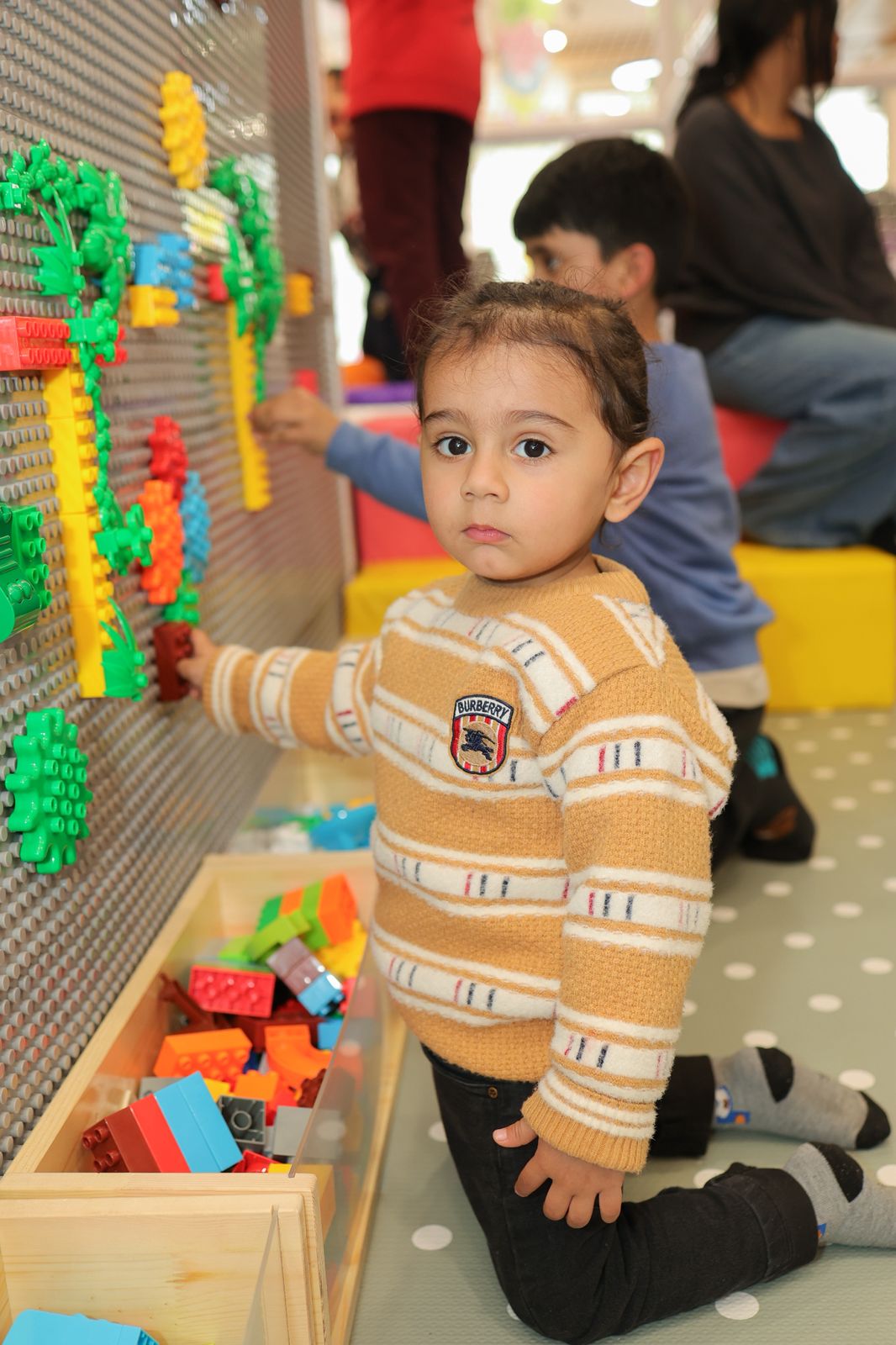 A young child plays with colorful building blocks on a wall, while other children and adults are in the background.