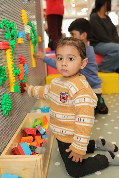 A young child plays with colorful building blocks on a wall, while other children and adults are in the background.