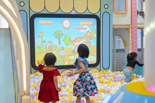 Two young girls play a projected game in a ball pit, with another child in the background.