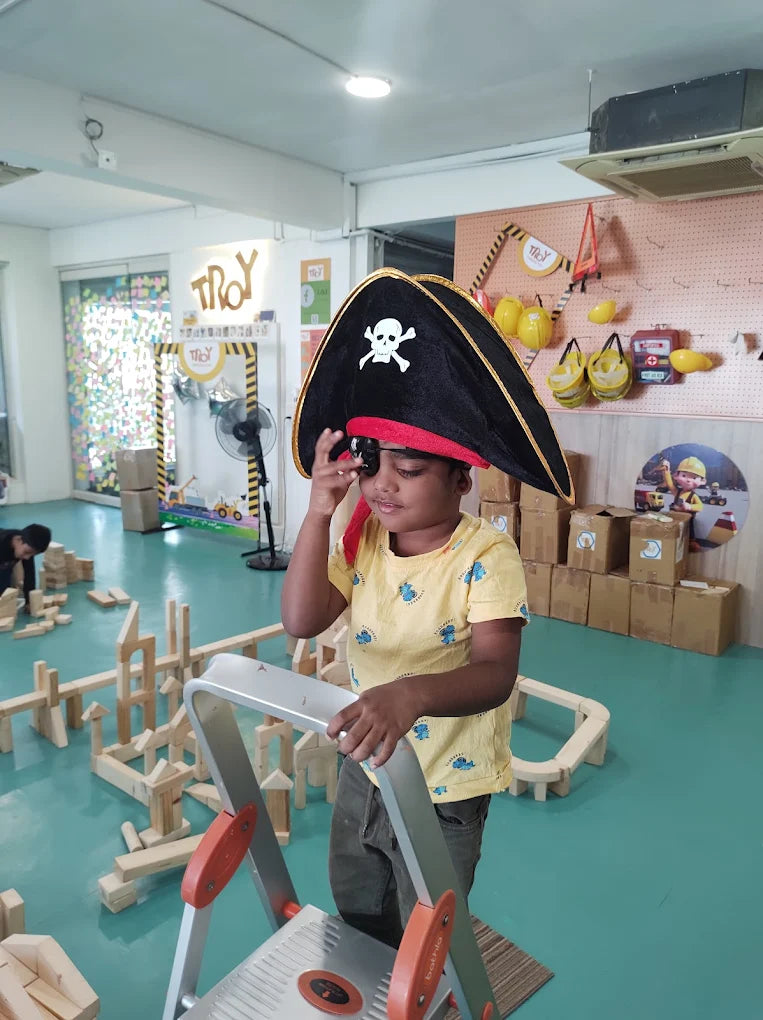 A young child dressed as a pirate stands on a step stool in a playroom filled with wooden blocks.
