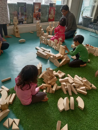 Children and an adult play with wooden blocks on a green carpet.