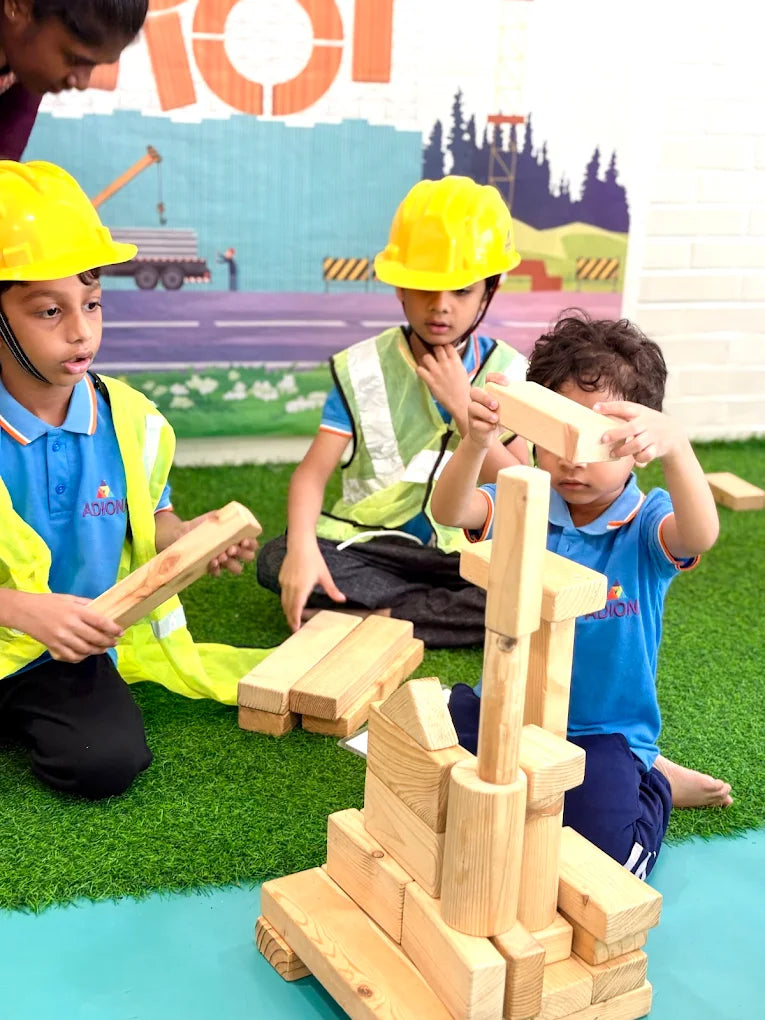 Young children in hard hats play with wooden blocks on a grassy area, building a structure.
