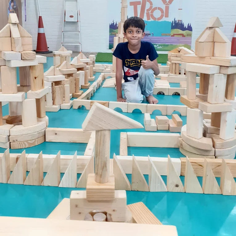 A young boy gives a thumbs-up while sitting amidst a large structure of wooden blocks.
