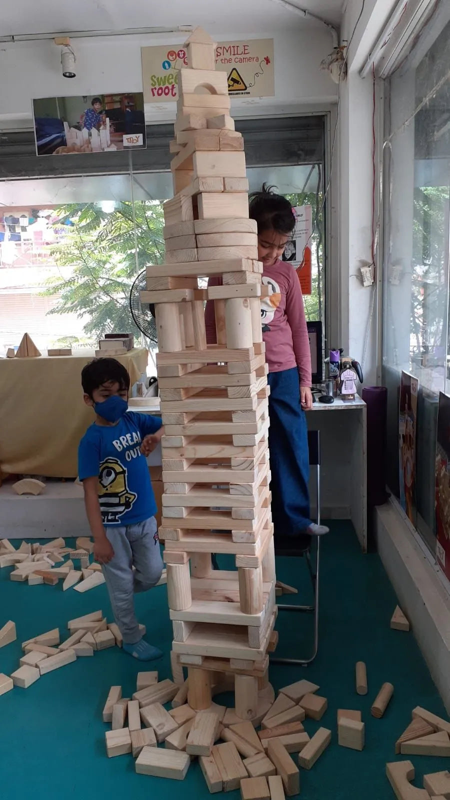 Two children build a tall tower of wooden blocks in a room with a window.