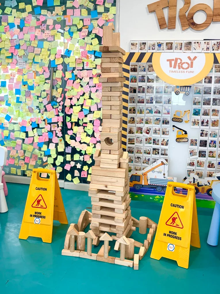 A tall Jenga tower stands in a room decorated with sticky notes and photos, with two "Work in Progress" signs in the foreground.