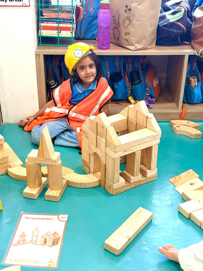 A young child in a hard hat and safety vest plays with wooden blocks, building a village.