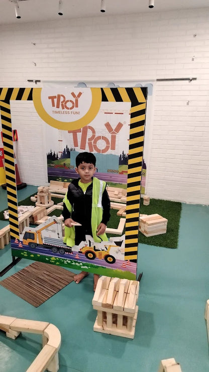 A young boy in a safety vest stands in a construction-themed play area with large wooden blocks.