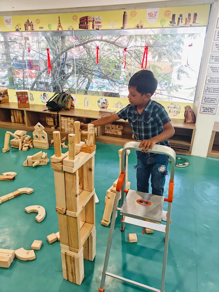 A young boy builds with wooden blocks in a play area, using a step stool to reach higher.