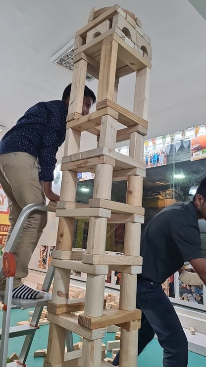 Two young men build a tall tower of wooden blocks, one on a ladder.