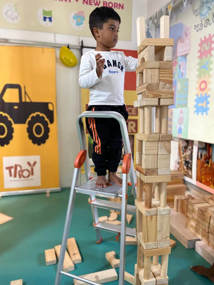A young boy stands on a ladder, building a tall tower of wooden blocks. He is in a room with colorful decorations and other blocks scattered around.