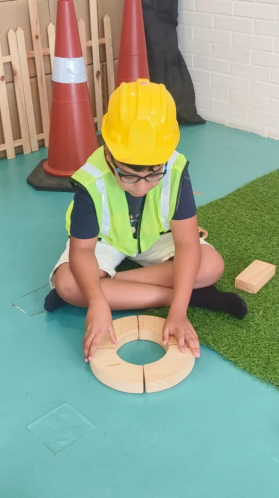 A young child in a hard hat and safety vest plays with wooden blocks on a blue floor next to a patch of artificial grass.