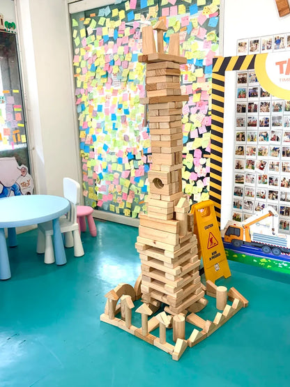 A tall tower of wooden blocks stands in a playroom, surrounded by colorful sticky notes on a glass wall.
