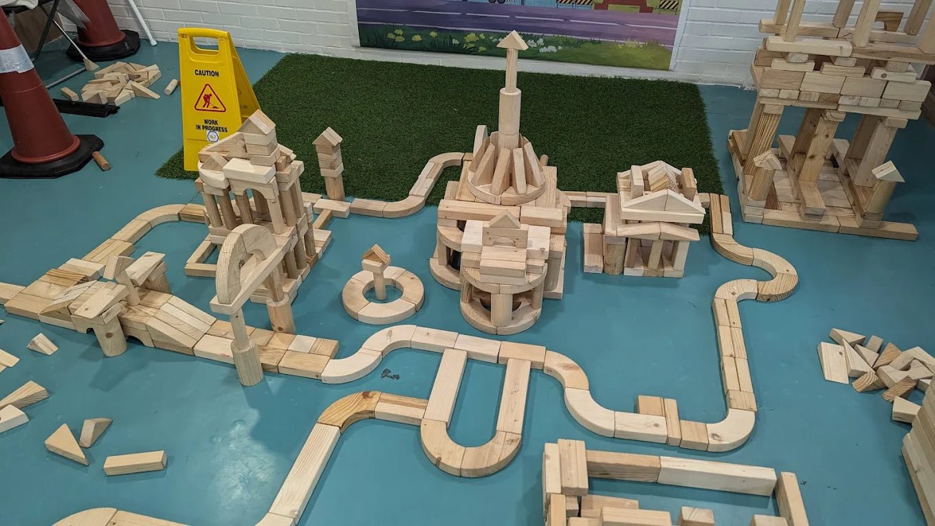 Children's wooden blocks are arranged in a complex structure on a blue floor, with a caution sign in the background.