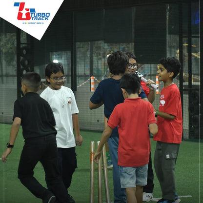 Young boys play cricket indoors on a turf field, with some holding a bat and others standing near wickets.